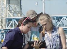 Three students looking at an iPad in front of the Portage Lake Lift Bridge.